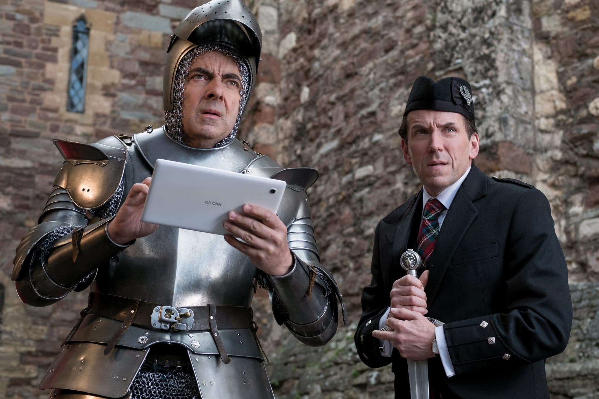 Johnny English (Rowan Atkinson) hält ein Tablet in seinen Händen während er eine Ritterrüstung trägt. Neben ihm steht sein Partner Rough (Ben Miller) in Uniform mit Schwert in den Händen.