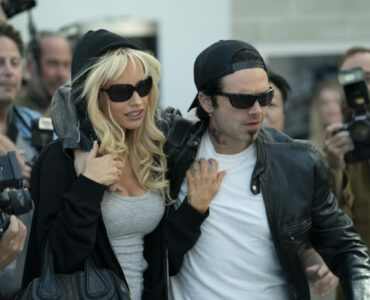 Pamela Anderson und Tommy Lee kämpfen sich durch eine Menge von Papparazzi. Beide sind dunkel gekleidet und tragen Sonnenbrillen - Pam & Tommy