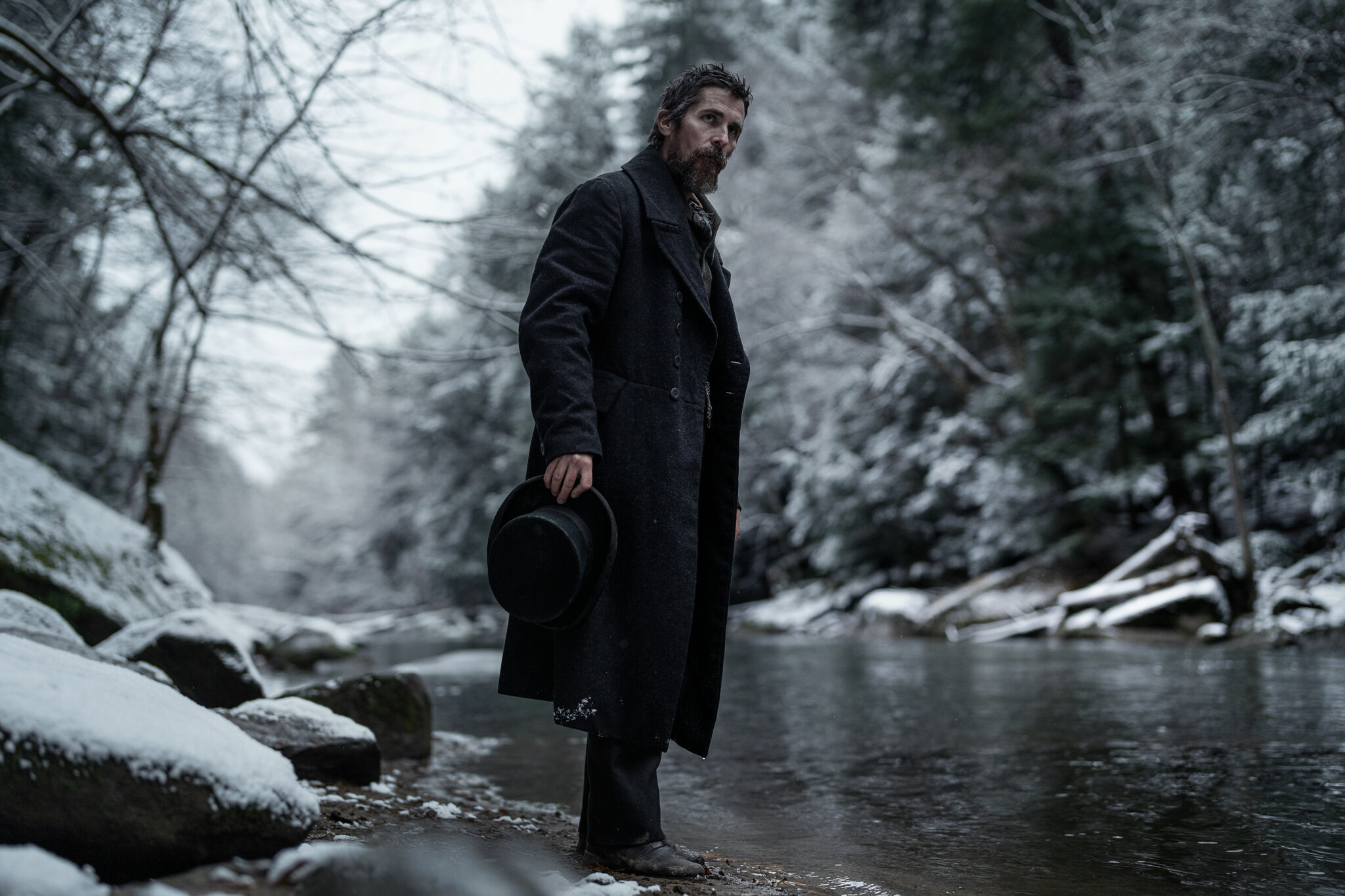 Christian Bale als Augustus Landor an einem winterlichen Fluss stehend mit schwarzem Mantel und Zylinder in der rechten Hand.