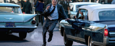Auf dem Bild sind Autos aus den 1960er Jahren zu sehen. Zwischen zweien rennt Harrison Ford in einem Anzug und Krawatte als Indiana Jones über die Straße. Es ist ein Szenenbild aus Indiana Jones und das Rad des Schicksals.