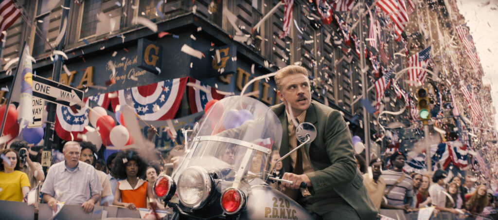 Im Hintergrund ist eine Parade in der US-Stadt New York zu erkennen. Im Vordergrund sitzt ein Mann mit Schnauzbart und Kurzhaarschnitt auf einem Motorrad. Es ist der Schauspieler Boyd Holbrook und es ist eine Szene aus Indiana Jones und das Rad des Schicksals.