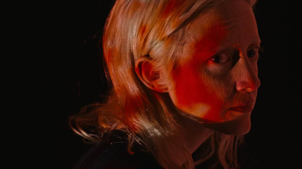 Andrea Riseborough, die Hauptdarstellerin von Possessor, schaut vom Neonlicht umhüllt provokant in die Kamera.