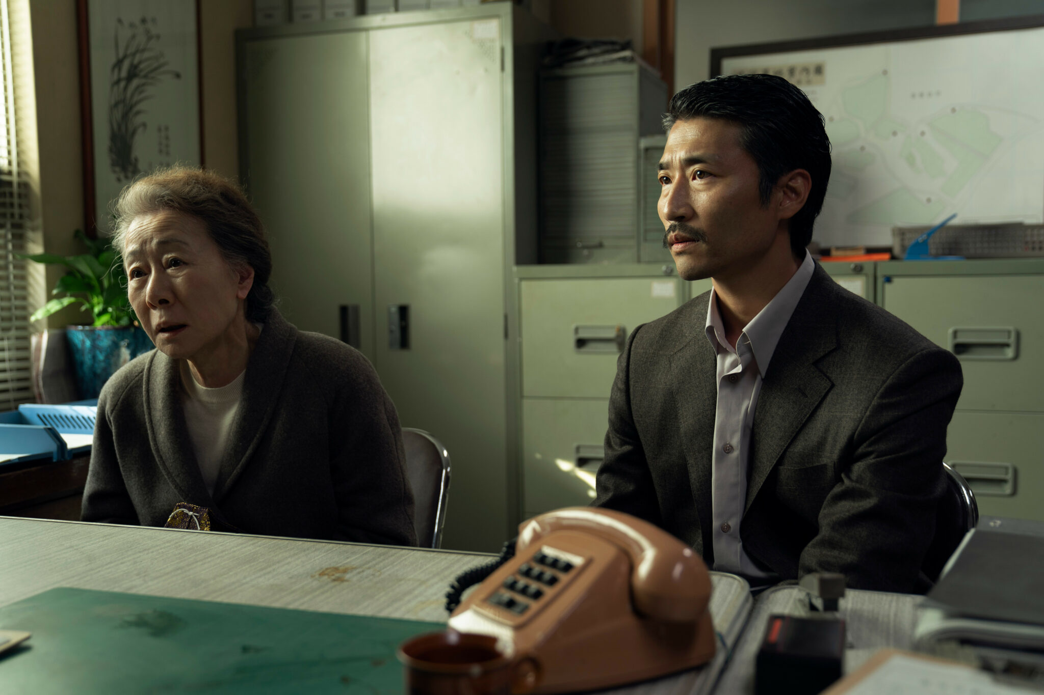 Zwei Koreaner sitzen hinter einem Schreibtisch auf dem ein rotes Telefon steht. im Hintergrund stehen Aktenschränke