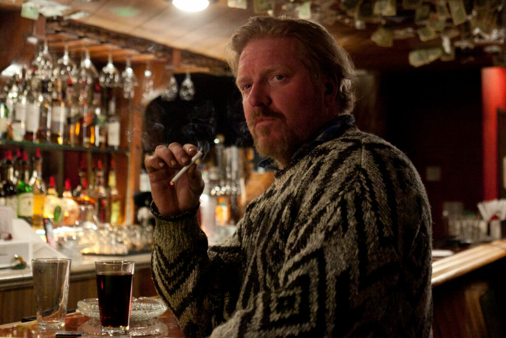 Im Film Pig Killer sitzt Jake Busey als Willy Pickman an der Bar in einem Pub und raucht eine Zigarette, vor sich ein Glas Whisky-Cola auf dem Tresen stehend.