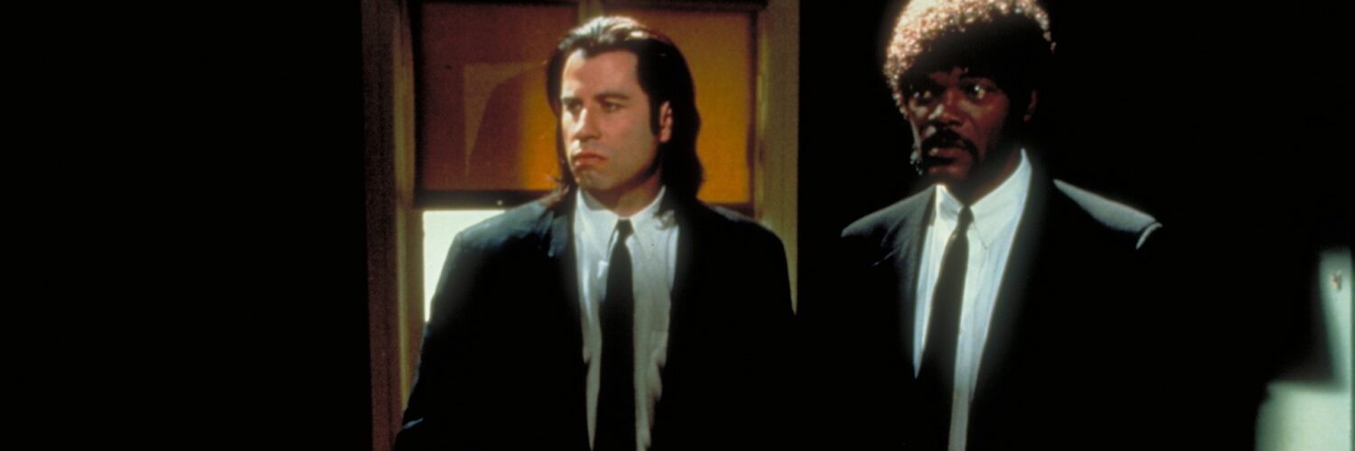Zwei Männer in schwarzen Anzügen, links ein weißer Mann mit dunklen langen Haaren, rechts daneben ein dunkelhäutiger Mann mit Bart und gelockten Haaren. Es sind die Schauspieler John Travolta und Samuel L. Jackson im Film Pulp Fiction.