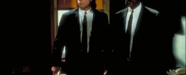 Zwei Männer in schwarzen Anzügen, links ein weißer Mann mit dunklen langen Haaren, rechts daneben ein dunkelhäutiger Mann mit Bart und gelockten Haaren. Es sind die Schauspieler John Travolta und Samuel L. Jackson im Film Pulp Fiction.