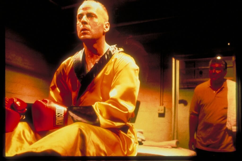 Ein glatzköpfiger Mann im gold-gelben Satin-Bademantel, rote Boxhandschuhe an den Händen. Es ist der Schauspieler Bruce Willis in einer Filmszene des Films Pulp Fiction.