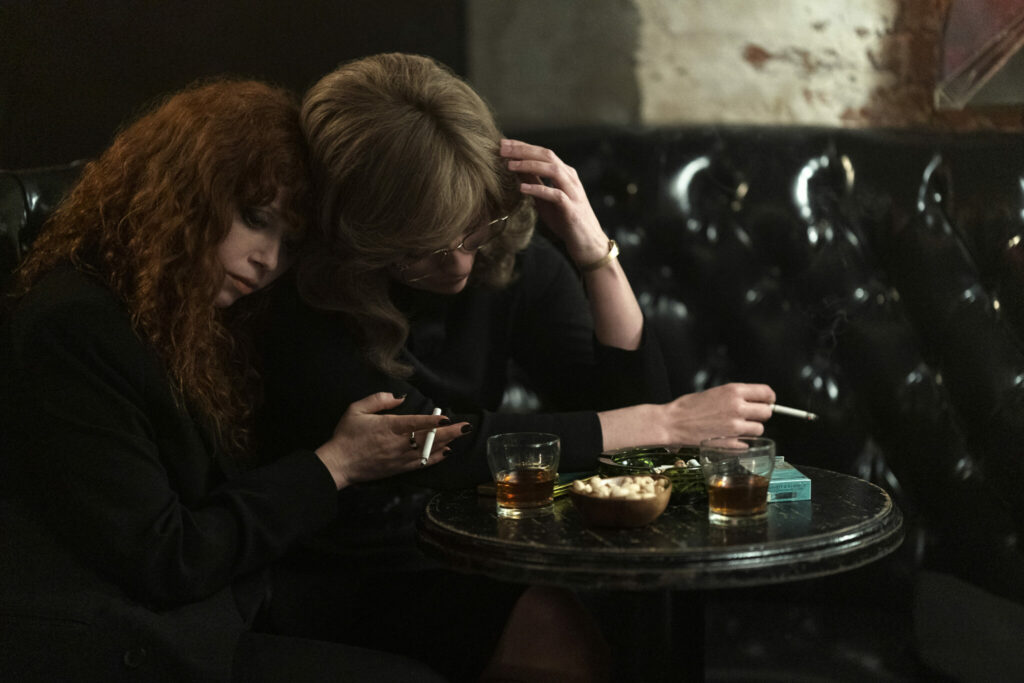 Natasha Lyonne klammert sich an Annie Murphy, die telefoniert. Dabei sitzen sie rauchend an einem Holztisch auf einem schwarzen Ledersofa - Matrjoschka Staffel 2.