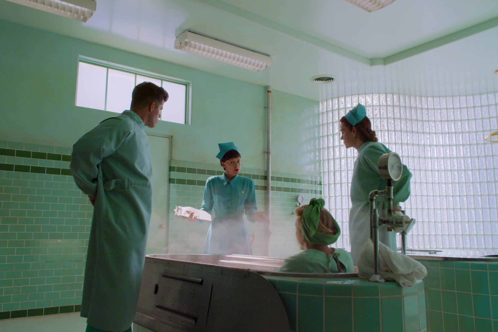 Huck (Charlie Carver) und die Schwestern Betsy Bucket (Judy Davis) und Mildred Ratched (Sarah Paulson) stehen in einem grünen Behandlungsraum um eine Wanne für eine Therapiemethode herum, in der schon eine Patientin platz genommen hat. 