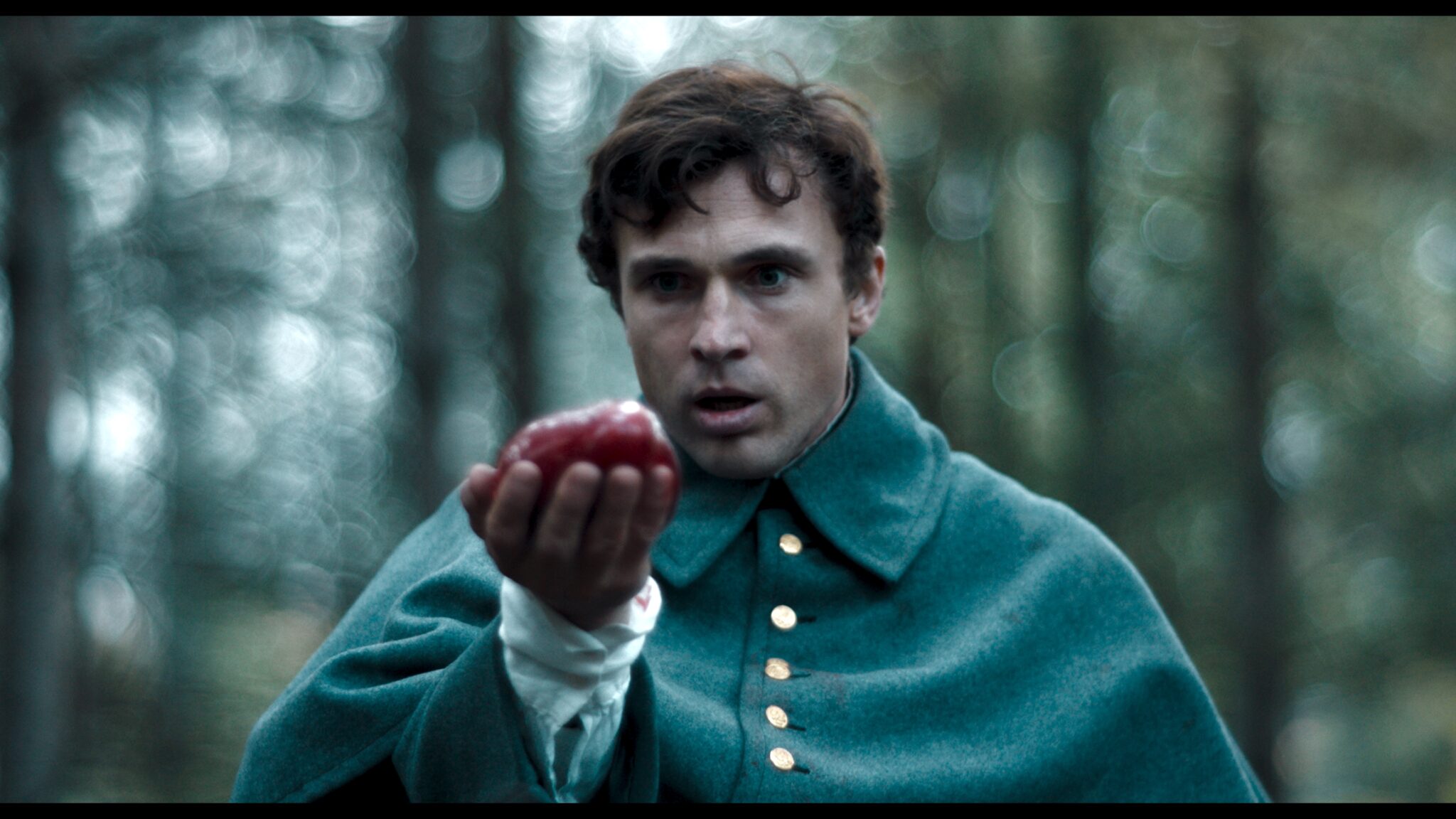 William Moseley steht, im grünen Umhang gewandet, im Wald und betrachtet erstaunt einen roten Apfel, den er vor sich in der Hand hält - Raven's Hollow