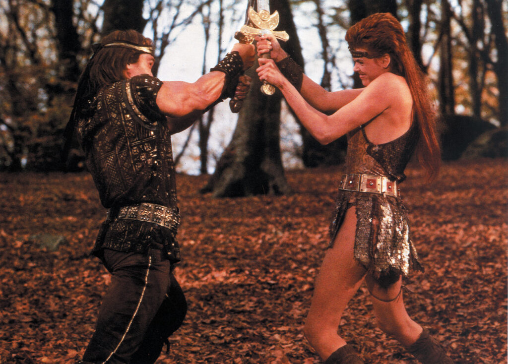 Ein mit Schwert kämpfender Arnold Schwarzenegger trägt bräunliche Kleidung, die nach Mittelalter aussieht. Gegen ihn kämpft Brigitte Nielsen, die einen Metallrock anhat. Eine Szene aus dem Film Red Sonja.