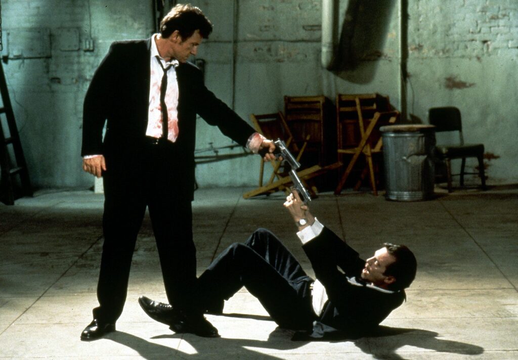 Zwei Männer, der rechts im Bild liegend, bedrohen sich gegenseitig mit einer Waffe. Links steht der Schauspieler Harvey Keitel und rechts liegt Tim Roth. Beide tragen einen schwarzen Anzug und ein weißes Hemd mit einigen blutigen Flecken. Es handelt sich um eine Szene aus dem Film Reservoir Dogs.