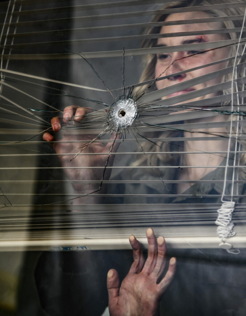 Zoe, gespielt von Isabel May, blickt in Run Hide Fight durch eine zerschossene Fensterscheibe nach draußen auf der Suche nach dem Schützen.