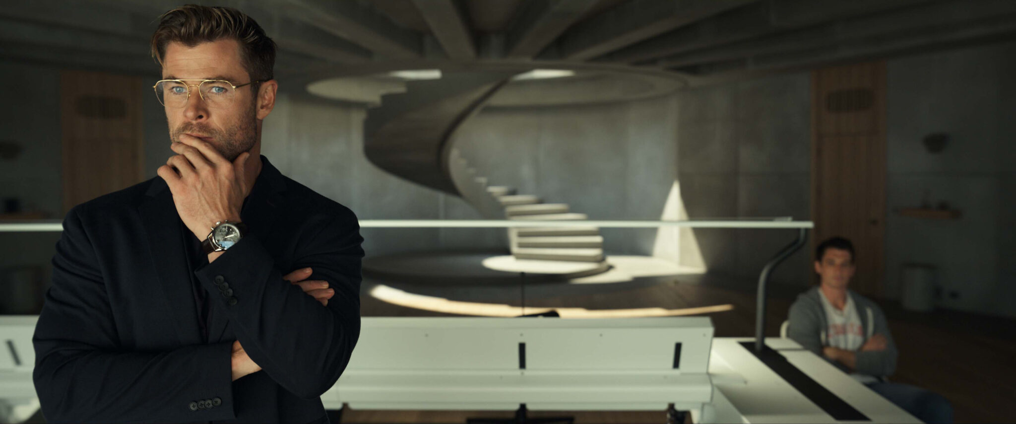 Chris Hemsworth in Der Spinnenkopf in einem Raum mit Betonwänden und einer Wendeltreppe in nachdenklicher Pose im Vordergrund.
