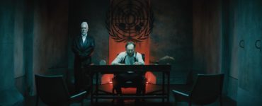 Der alte Präsident der Welt, gespielt von Mark Ashworth, sitzt in einem düsteren Raum an einem Schreibtisch vor einer rot erleuchteten Nische, flankiert von einem Wächter.
