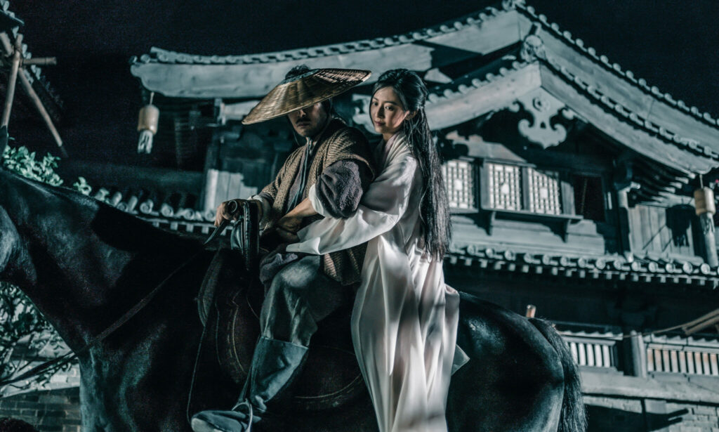 Donnie Yen, den Bambushut tief ins Gesicht gezogen, sitzt mit Yuqi Chen auf einem Pferd - Sakra