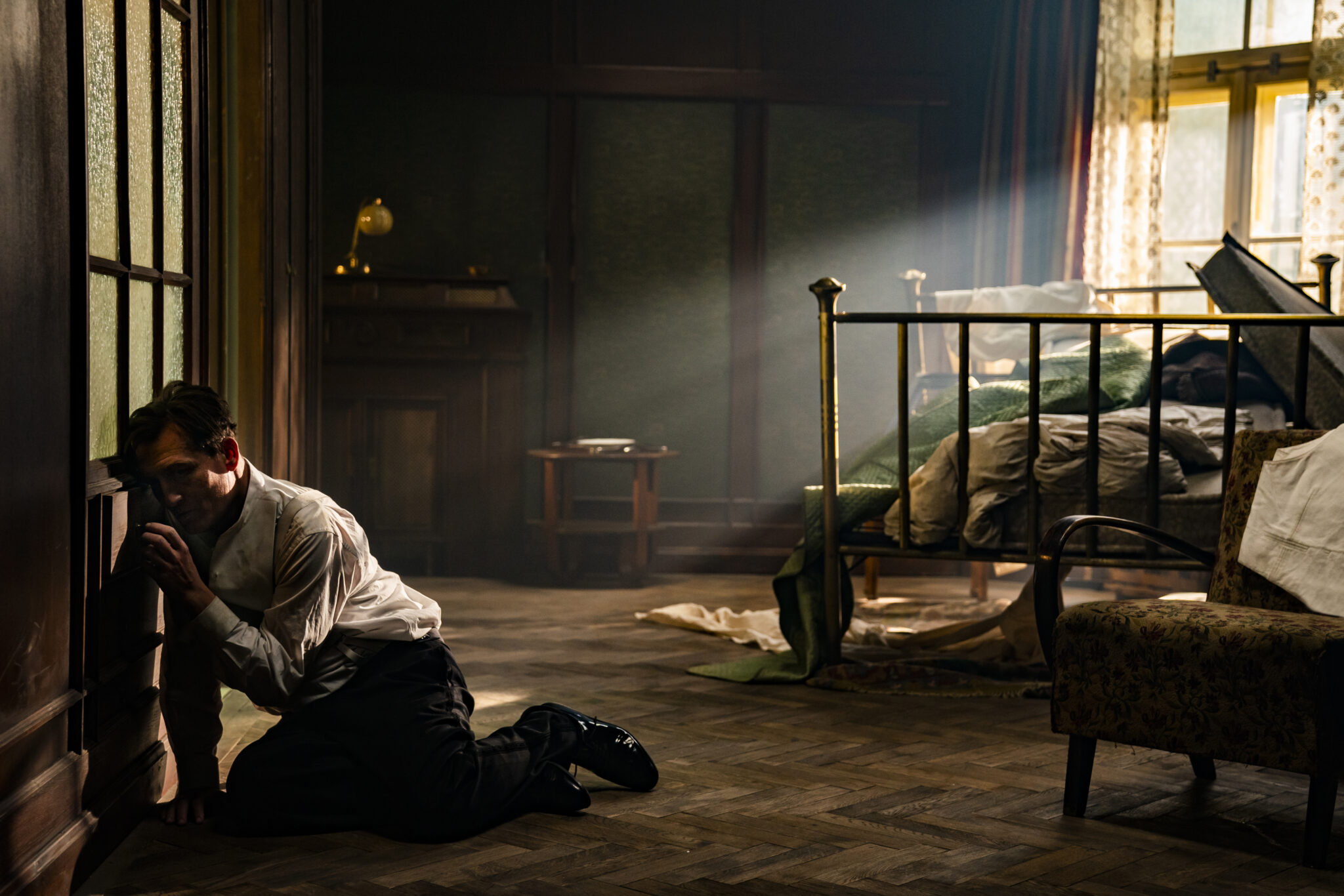 Bartok (Oliver Masucci) lehnt links an einer Tür kauernd. Von rechts hinten scheint die Sonne durch ein Fenster. Rechts im Raum stehen ein Sessel im Vordergrund und ein zerwülltes Bett dahinter. 