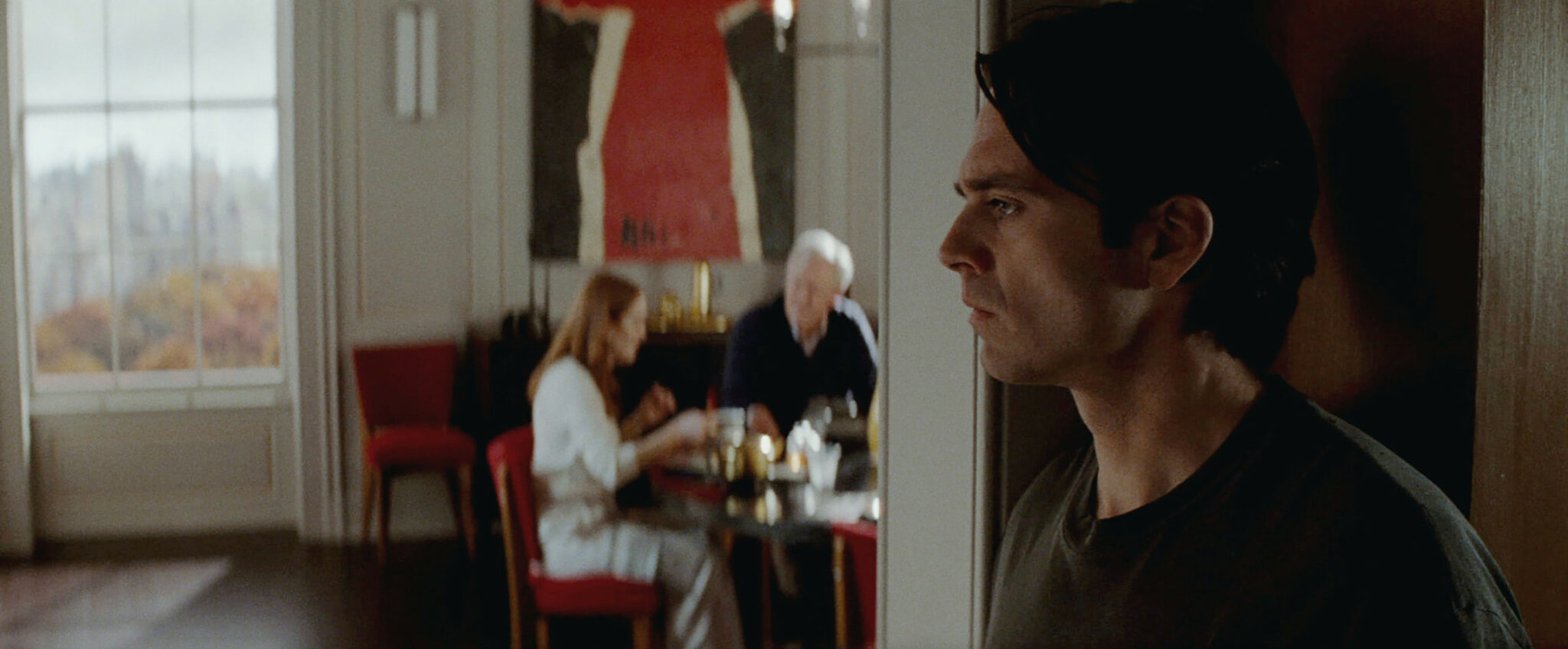 Sebastian Stan im Türrahmen stehend im Vordergrund, hinten Julianne Moore und John Lithgow an einem kleinen Tisch sitzend. Sharper