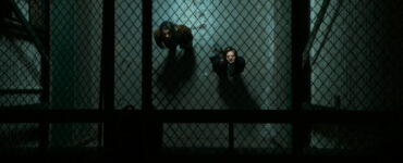 Ein Blick von oben durch ein Gitter auf zwei Personen in einem Gang, die nach oben blicken.