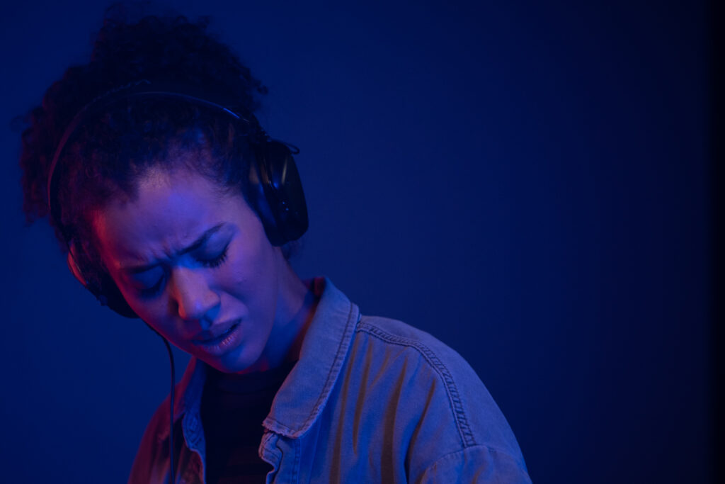 Jasmin Savoy Brown steht als Alexis im Film Sound of Violence vor einer blauen Wand, trägt Kopfhörer und lauscht konzentriert der Musik