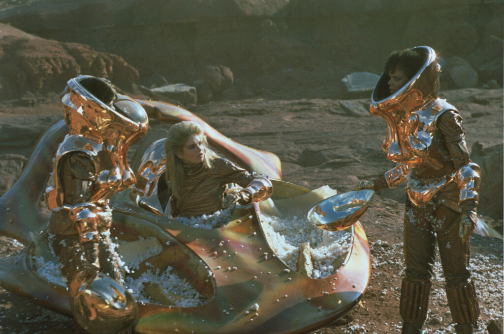 Drei Frauen in spiegelnd metallenen Anzügen steigen aus dem gestrandeten, ebenso spiegelnd metallenen, Gefährt - Spacehunter - Jäger im All.