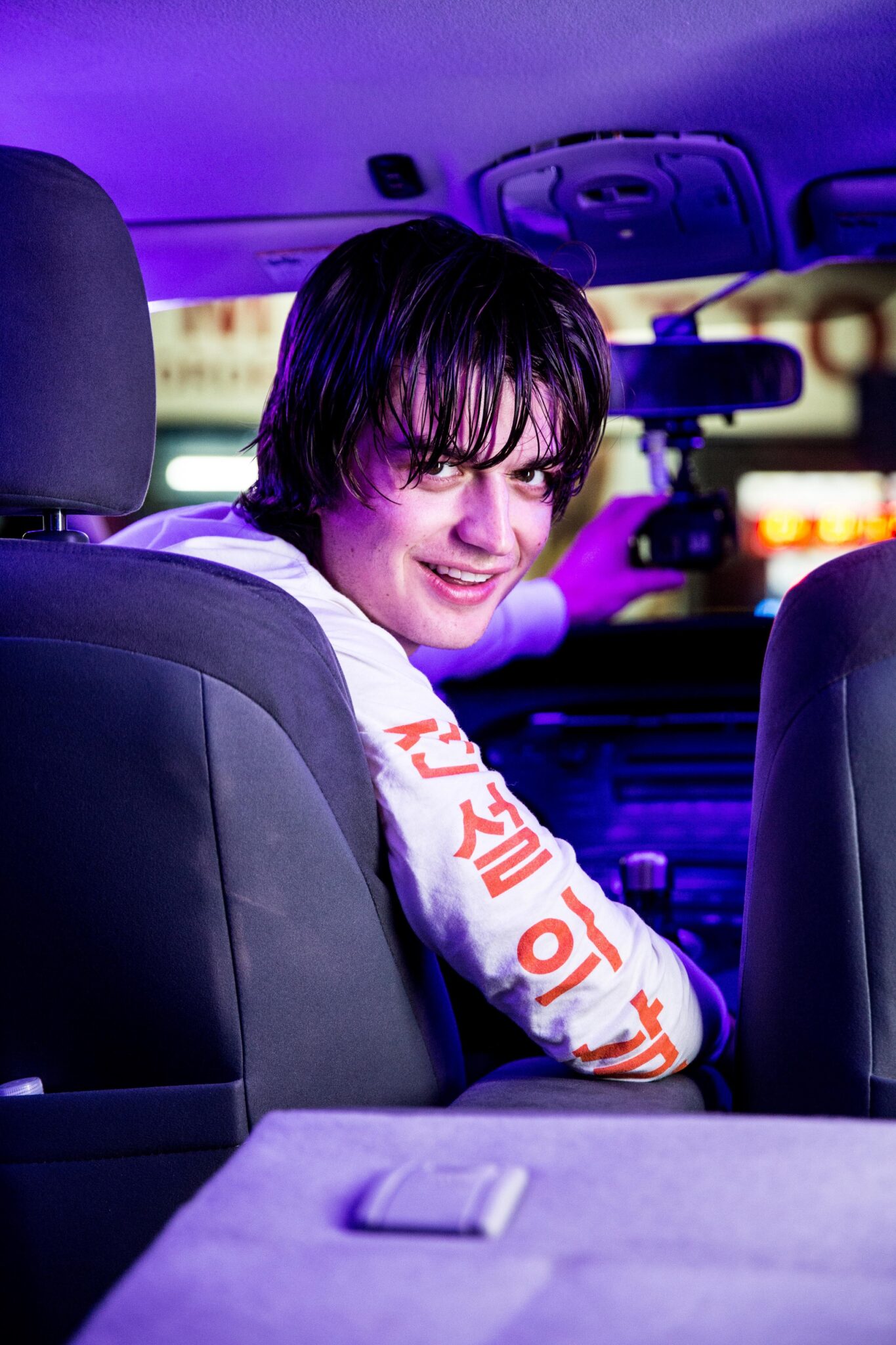 Joe Keery als Kurt Kunkle im Film Spree - Alles für die Klicks in seinem Privattaxi auf dem Fahrersitz wie er mit der linken Hand die Dashcam richtet und verschmitzt über die rechte Schulter auf den Rücksitz in die Kamera grinst.