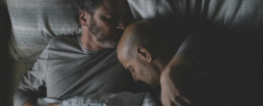 Sam (Colin Firth) und Tusker (Stanley Tucci) liegen gemeinsam im Bett. Sam hat den Arm um Tusker gelegt und küsst ihn auf den Kopf.