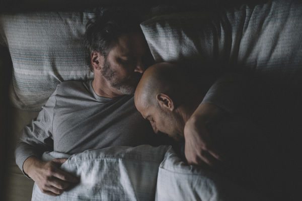 Sam (Colin Firth) und Tusker (Stanley Tucci) liegen gemeinsam im Bett. Sam hat den Arm um Tusker gelegt und küsst ihn auf den Kopf.