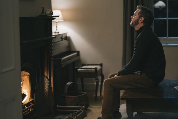 Sam (Colin Firth) sitzt auf einem Tisch und schaut in die Leere. Im Vordergrund ist ein Kamin mit brennendem Feuer, im Hintergrund ein Klavier.