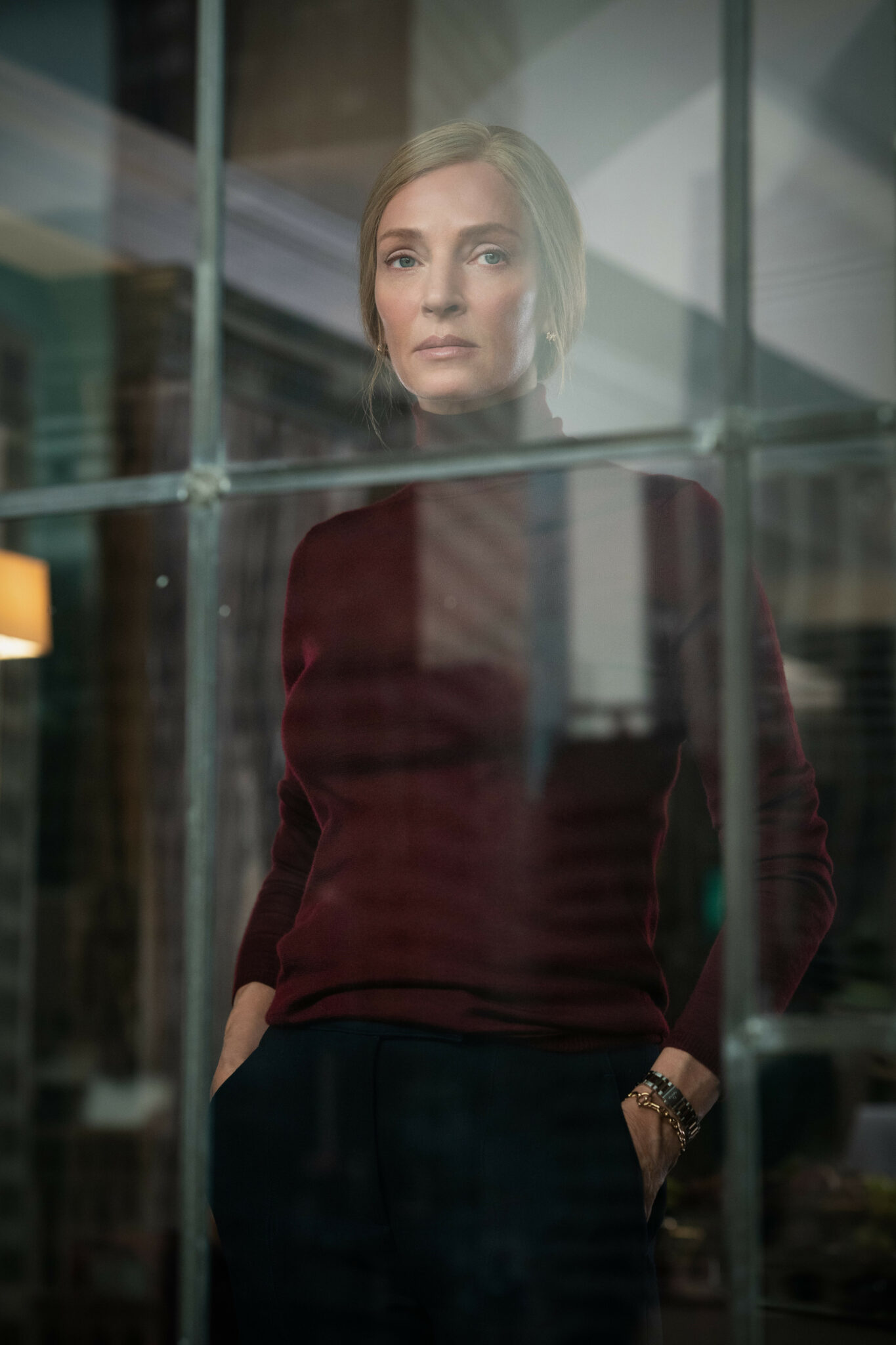 Uma Thurman hinter einer Fensterscheibe mit den Händen in den Hosentaschen und einem roten Rollkragenpullover. Suspicion