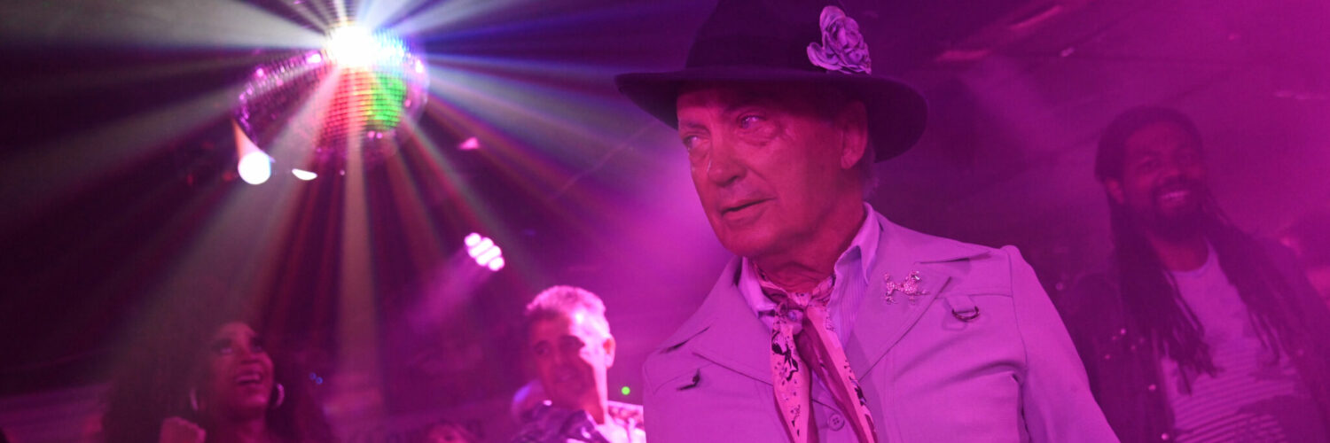 Udo Kier steht im lila getünchten Licht mit anderen Leute auf der Tanzfläche, über ihnen blitzt eine Discokugel - Swan Song