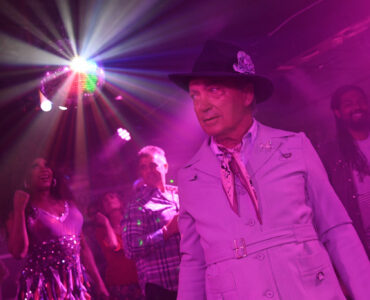 Udo Kier steht im lila getünchten Licht mit anderen Leute auf der Tanzfläche, über ihnen blitzt eine Discokugel - Swan Song