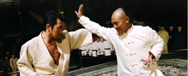In der Amerikanischen Einstellungsgröße stehen sich Huo Yuanjia auf der rechten Seite und sein Gegner gegenüber und bekämpfen sich. Im Bildhintergrund schauen Kampfkünstler dem Geschehen zu.