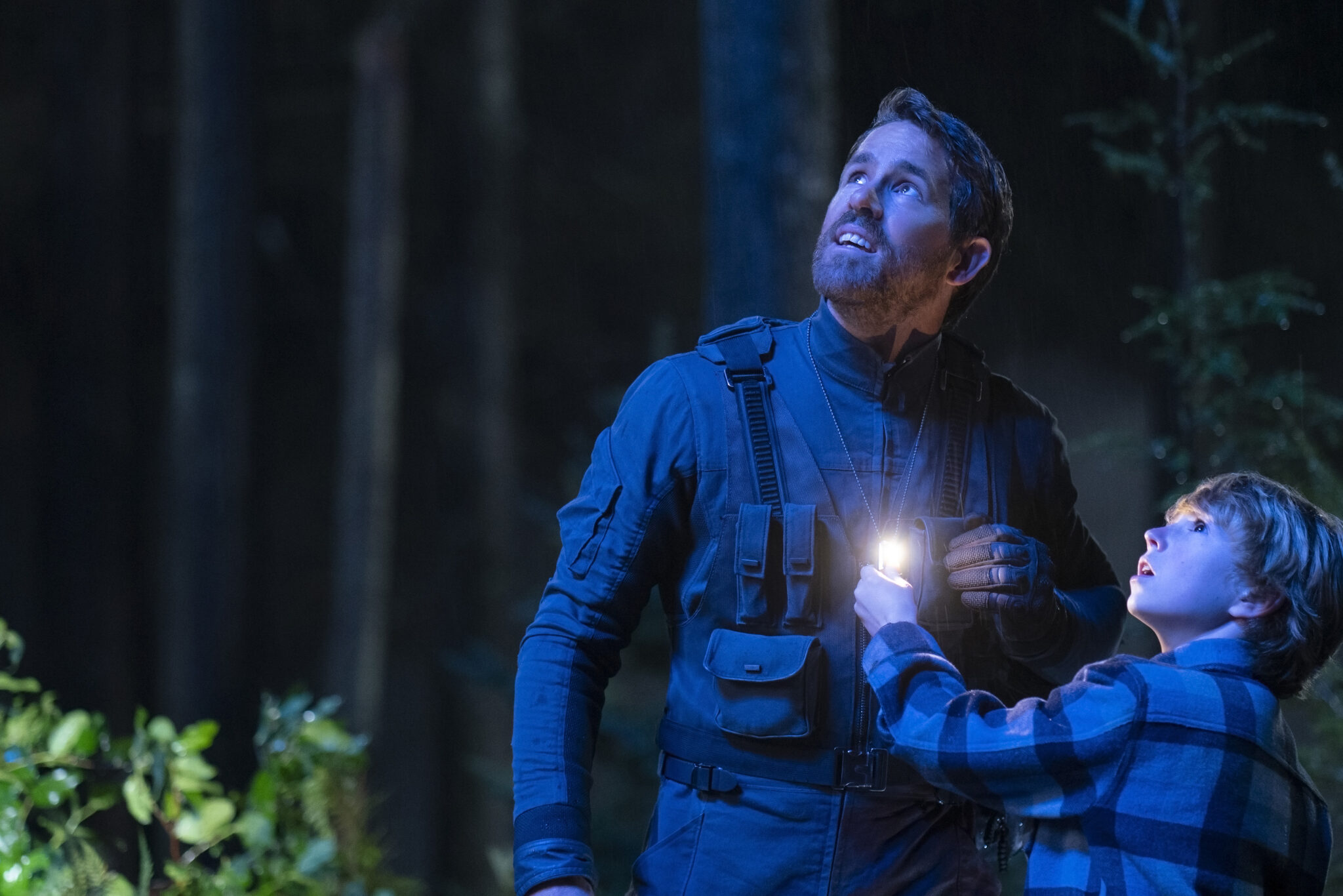 Ryan Reynolds neben einem kleinen Jungen im dunklen Wald blau angestrahlt. Beide blicken Richtung Himmel.