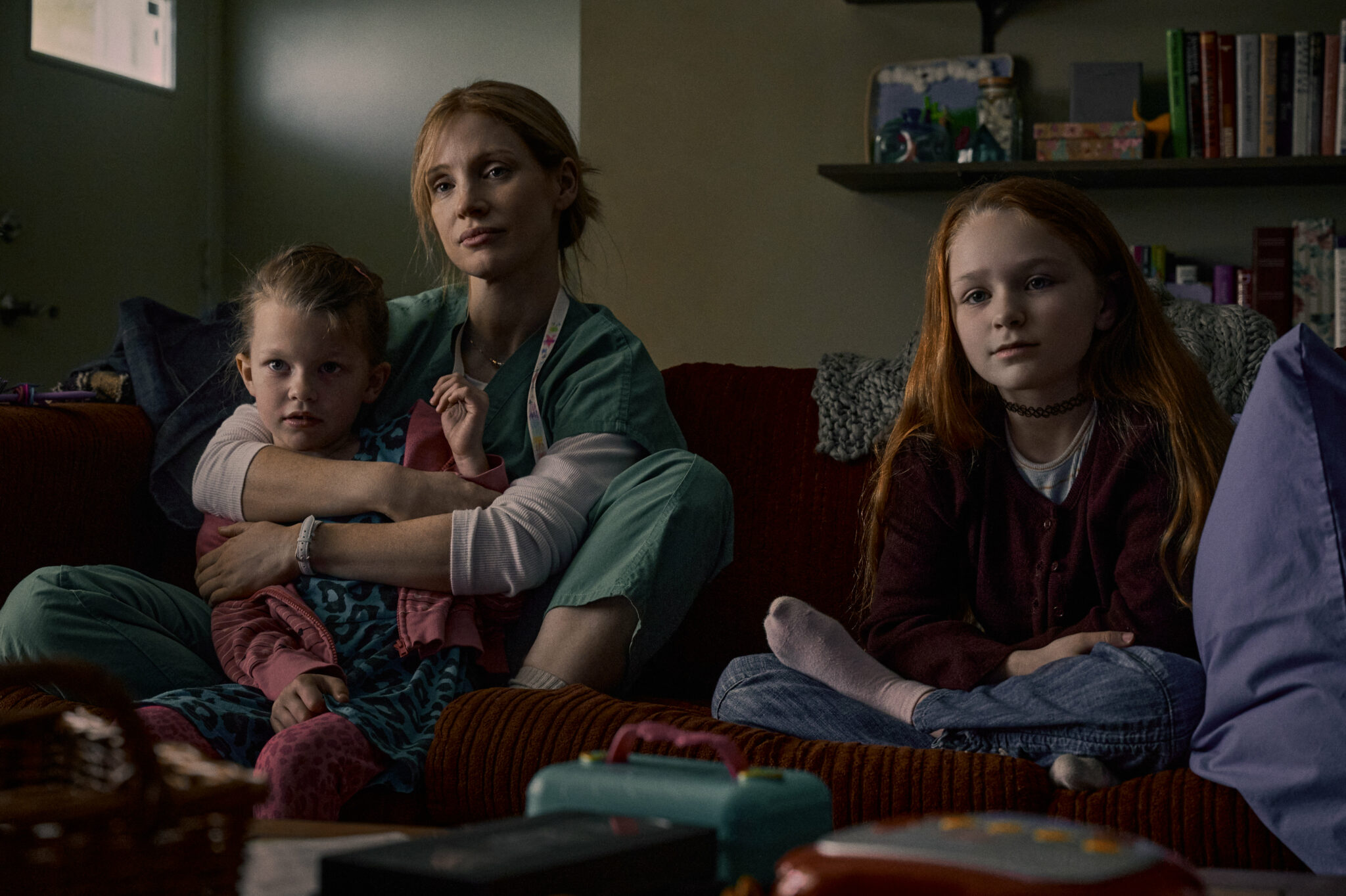 Jessica Chastain sitzt mit zwei Mädchen auf einer Couch. Sie trägt ein Schwestern-Outfit. Im Hintergrund ein Bücherregal. The Good Nurse
