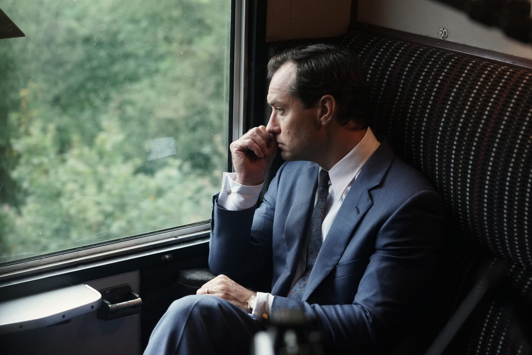 Jude Law sitzt in einem Zug. Er trägt Anzug und blickt gedankenverloren aus dem Fenster. The Nest