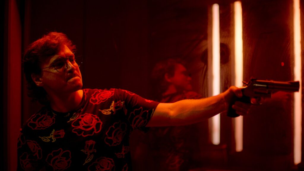Emile Hirsch als Alex zielt im Film The Price We Pay mit seinem Revolver in der linken Hand in einen rötlich ausgeleuchteten Raum