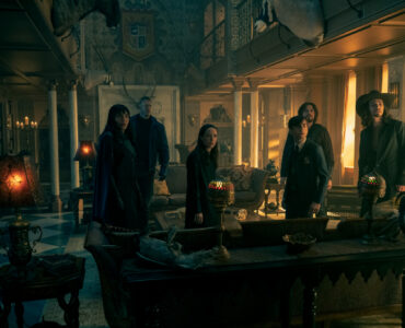 Die Geschwister der Umbrella Academy in einem verdunkelten Raum hinter einem Schreibtisch mit erstaunten Mienen.