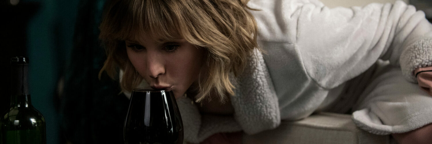 Kristen Bell beugt sich stehend über ein volles Weinglas und trinkt ab.