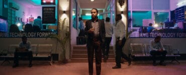 Balram steht mit einem Smartphone in der Hand vor einem neonbeleuchteten Büro. Er trägt dunkle Kleidung während Leute im Hintergrund helle Hemden tragen.
