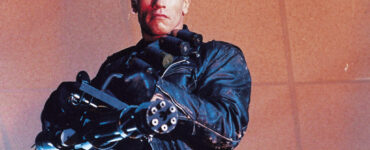 Ein Mann in Lederkleidung hält eine Minigun in unbekannte Richtung. Erkennbar ist es der Schauspieler Arnold Schwarzenegger in Terminator 2: Tag der Abrechnung