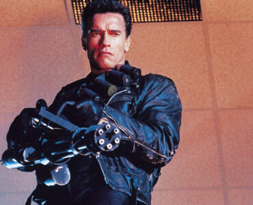 Ein Mann in Lederkleidung hält eine Minigun in unbekannte Richtung. Erkennbar ist es der Schauspieler Arnold Schwarzenegger in Terminator 2: Tag der Abrechnung