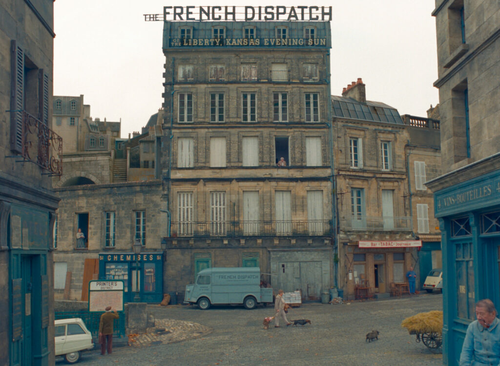 Die Redaktion des THE FRENCH DISPATCH haus in einem kleinen Hochhaus, im französischen Stil, vor dem ein blauer Lieferwagen parkt.
