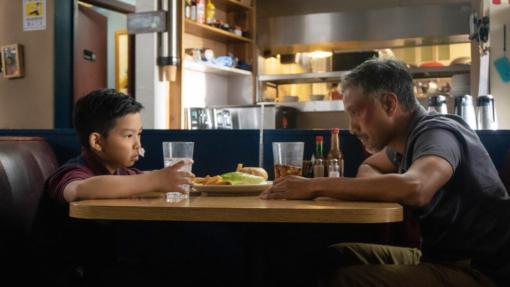 Danny sitzt mit seinem enttäuscht dreinblickenden Sohn bei Pommes, Burger und Limo in einem Bistro - The Paper Tigers.