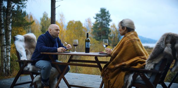 Lisa und Lars sitzen mit einem Glas Rotwein an einem Tisch. Im Hintergrund eine malerische Waldlandschaft. 