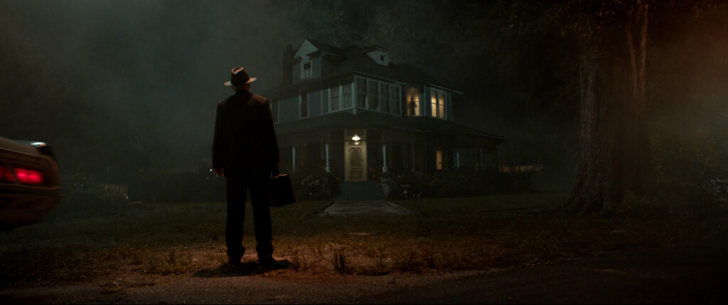 Es ist Nacht und ein Mann mit Hut und einer Tasche steht vor einem unheimlichen Landhaus