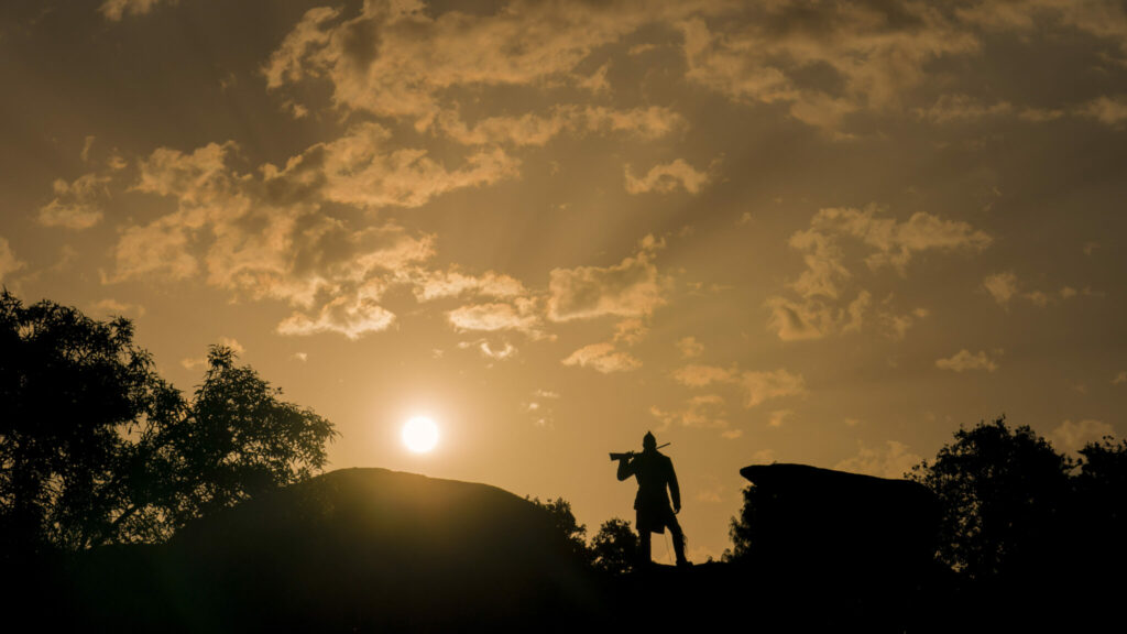 Eli Whipp, gespielt von Chaske Spencer, steht im Gegenlicht auf einem Hügel, sein Gewehr kampfbereit quer über seine Schultern gelegt.