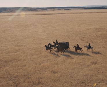 Eine Kutsche fährt über weites Land begleitet von mehreren Reitern.