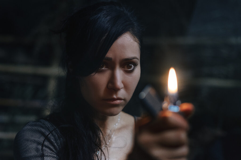 Cristina schaut im Dunkeln mit vor dem Gesicht erhobenen Feuerzeug in einer Nahaufnahme entschlossen Richtung Kamera | The Old Ways