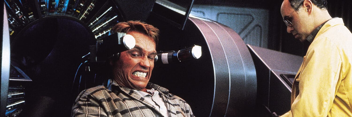 Douglas Quaid (Arnold Schwarzenegger) kämpft in Total Recall um seine Erinnerungen. Auf dem Bild sitzt Quaid gefesselt an einer Maschine. Sein Blick verrät, dass es schmerzhaft sein muss, oder zumindest gegen seinen Willen, denn er beisst sich sichtbar auf die Zähne. Zu seiner Linken steht noch ein Mann mit Halbglatze und Brille in einem gelben Kittel, der die Maschine scheinbar bedient.